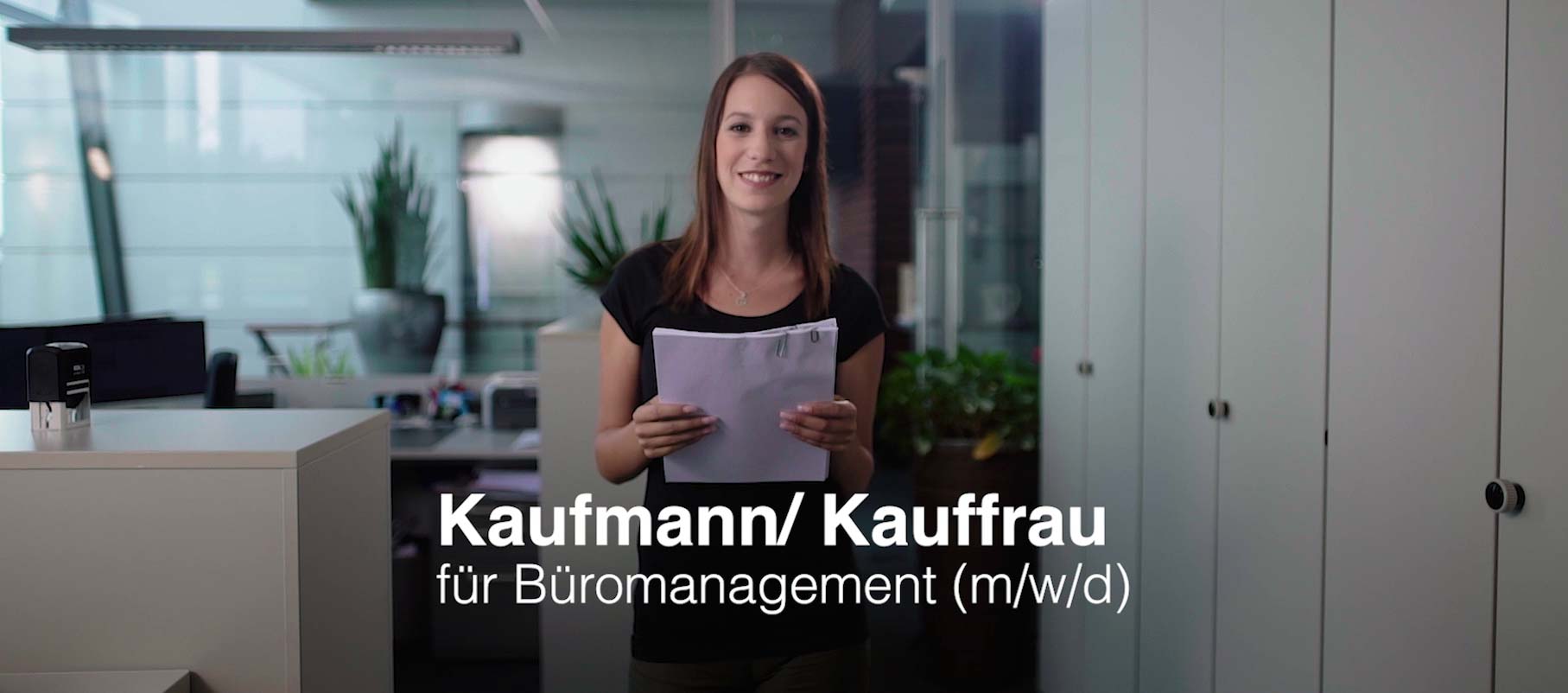 Junge Dame, lächelnd mit Akte in den Händen im Vordergrnd. Büro im Hintergrund. Aufschrift: "Kaufmann/ Kauffrau für Büromanagement (m/w/d)".