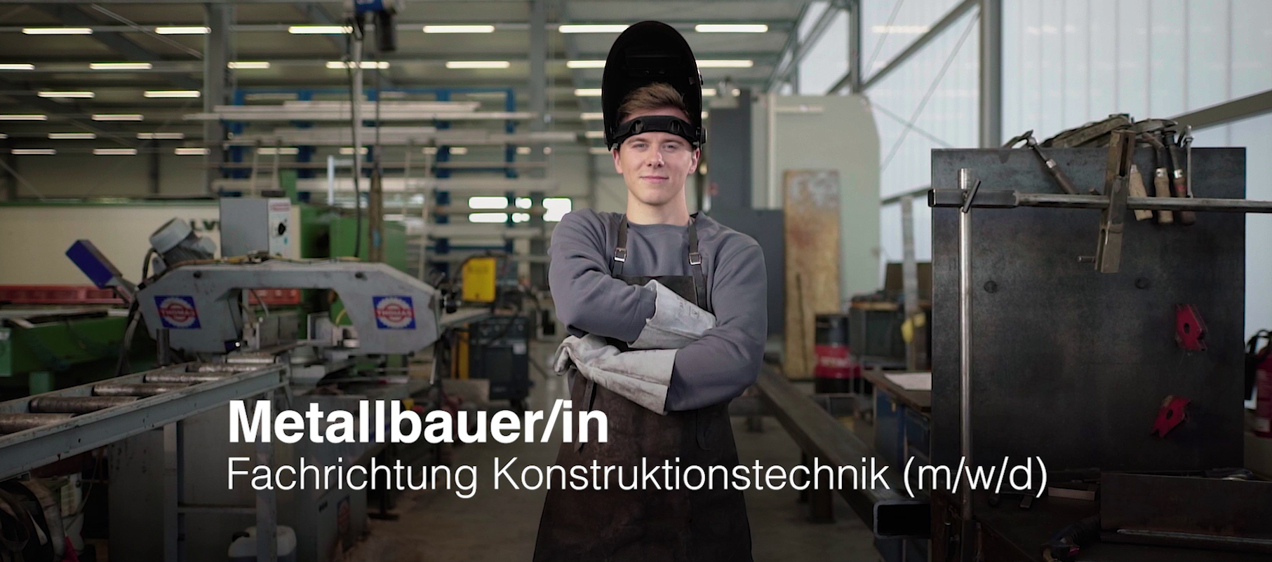 Jungermann mit verschränkten Armen im Vordergrund. Produktionshalle im Hintergrund. Aufschrift: "Metallbauer/in Fachrichtung Konstruktionstechnik (m/w/d)